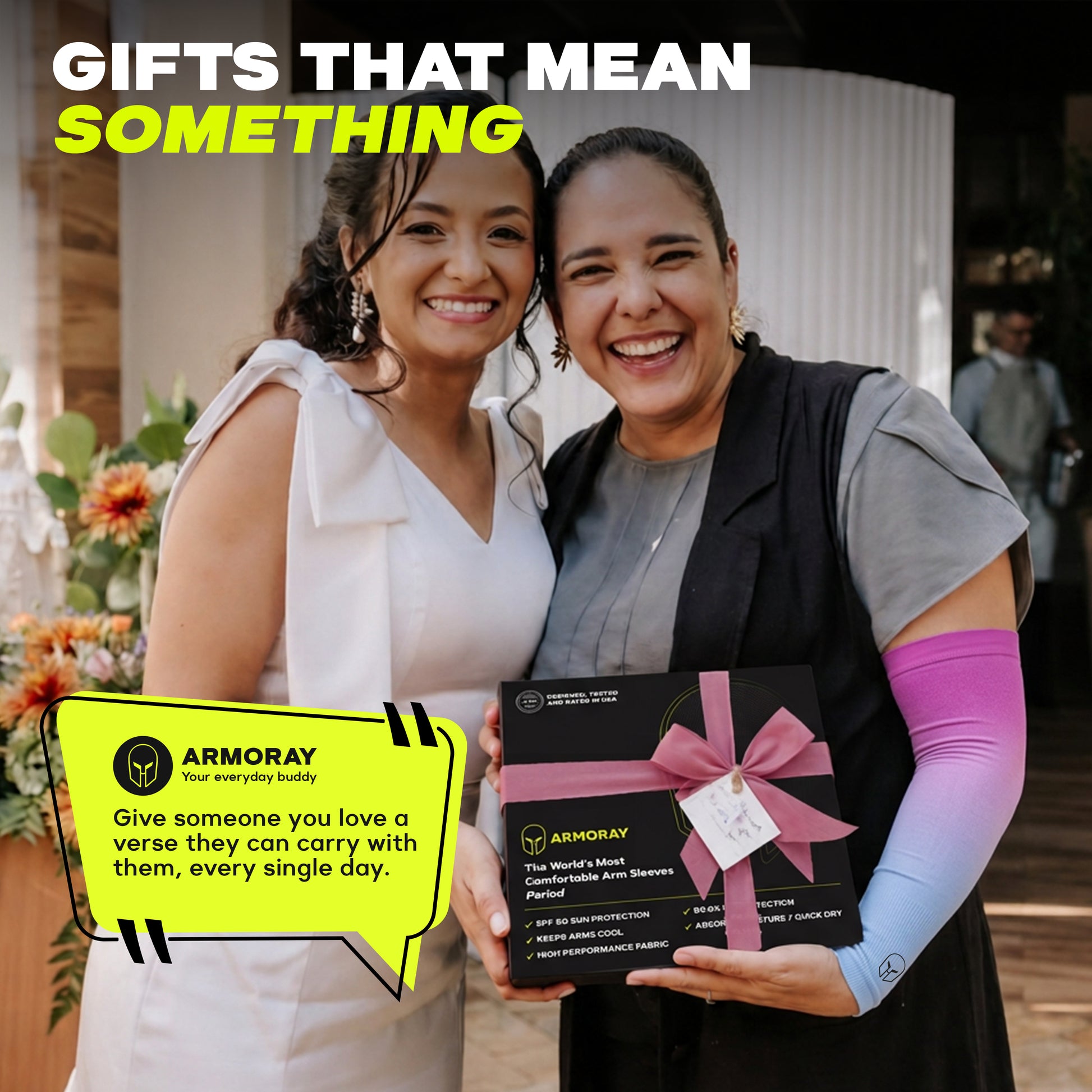 Two women stand smiling, one holding a black gift box with a pink ribbon and a yellow card, in a warmly lit indoor setting adorned with flowers. Overlaid text highlights the joy of meaningful gifts, such as the ARMORAY Ombré Collection—UPF 50+ arm sleeves that offer both stylish flair and reliable UV protection.