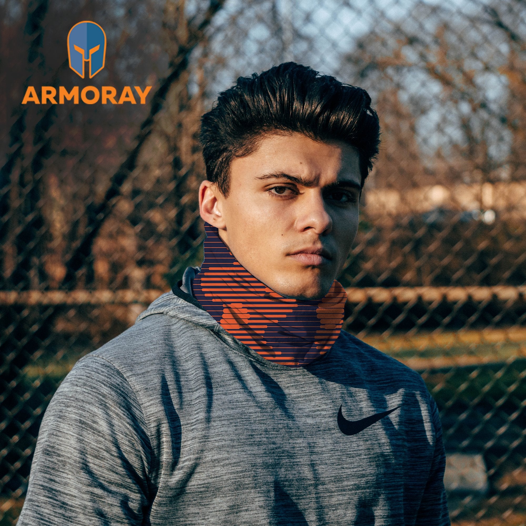 A young man outdoors wears a gray Nike hoodie and an Armoray® Bandanas — Designed for the Outdoors neck gaiter from ARMORAY, with the ARMORAY logo in the top left corner against a blurred fence background. Engineered for durability and sweat resistance, this rugged bandana is ideal for active lifestyles.