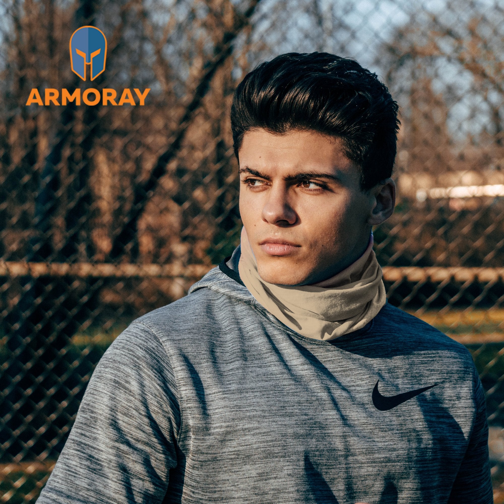 A young man in athletic wear and a neck gaiter stands outdoors by a chain-link fence. Sporting our Armoray® Bandanas — Designed for the Outdoors, he’s ready for anything. The ARMORAY logo appears in the upper left corner.